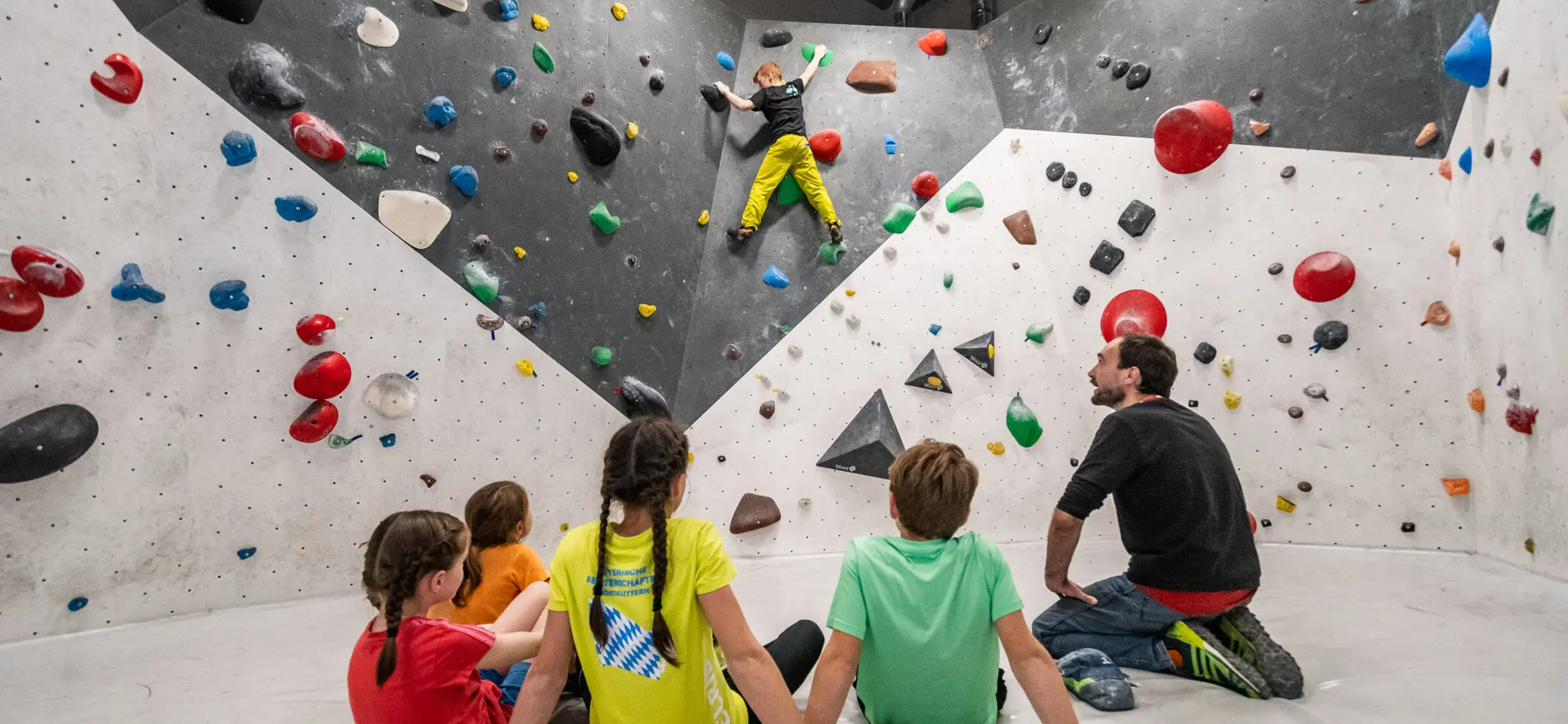 Eine Gruppe von Kindern sitzt in der DAV Kletterhalle Augsburg. Ein Kind bouldert an der Wand. Ferdinand Triller, Betriebsführer des DAV Kletterzentrum Augsburg, erklärt den Kids, wie sie am besten über schwierige Boulder kommen. | © Eckart Matthäus
